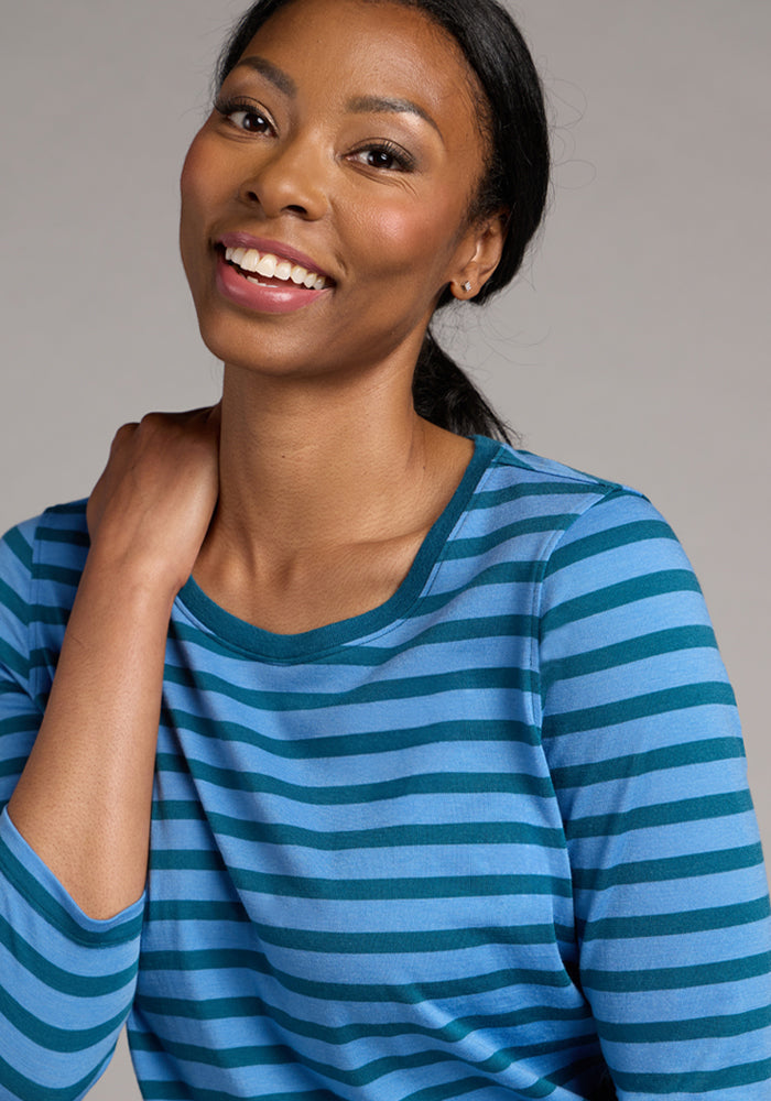 A smiling woman with long dark hair models the Woolx Jenny 3/4 Sleeve Crew Neck in Coastal Stripe, posing with one hand near her neck against a plain background.