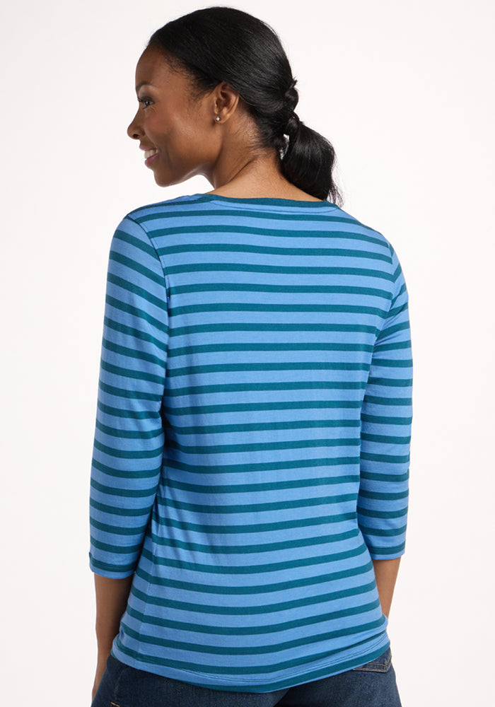 A woman with a low ponytail is seen from behind, wearing the Woolx Jenny 3/4 Sleeve Crew Neck in Coastal Stripe—a blue and turquoise lightweight Merino top—against a plain, light background.