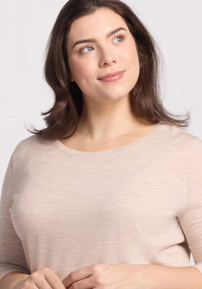 A woman with long, dark hair looks up and smiles softly, wearing a Jenny 3/4 Sleeve Crew Neck - Cream Heather by Woolx. The background is plain and light-colored.