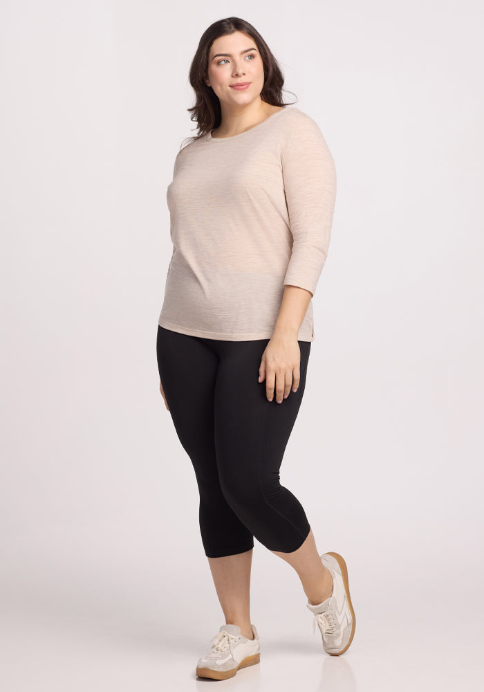 A woman with long dark hair smiles, wearing the Woolx Jenny 3/4 Sleeve Crew Neck in Cream Heather. She stands against a white background, paired with black leggings and white sneakers featuring beige soles.
