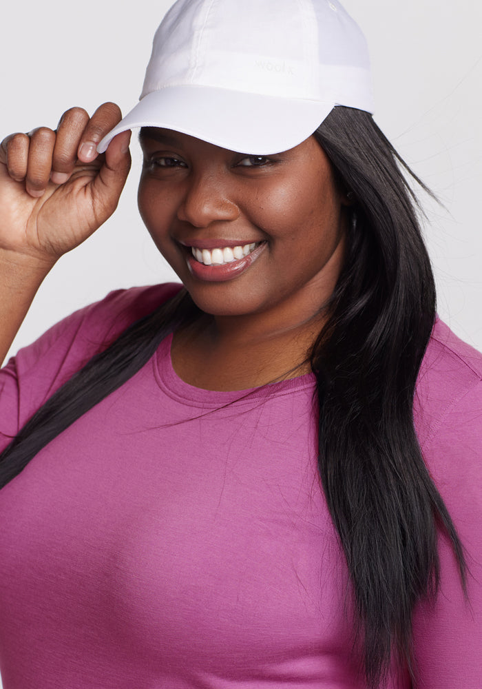 Smiling woman in a Woolx Jenny 3/4 Sleeve Crew Neck - Purple Wine, tilting her white cap with her right hand.
