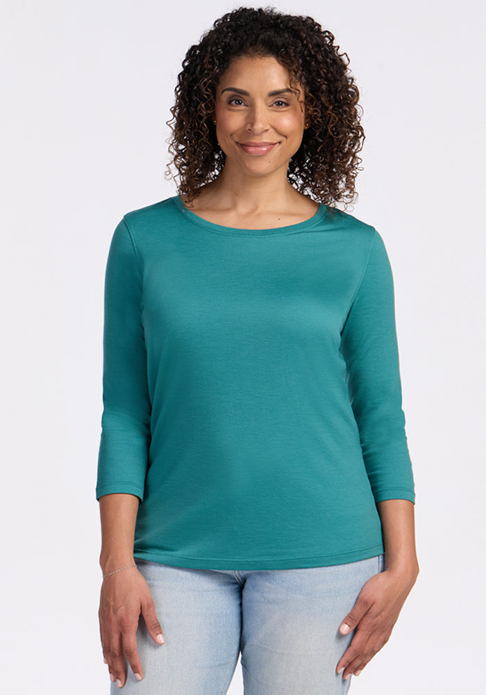 A woman with curly hair smiles in a Woolx Jenny 3/4 Sleeve Crew Neck - Surfside and light jeans against a plain background.
