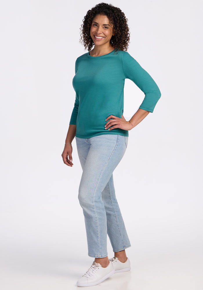 Woman with curly hair in a Woolx Jenny 3/4 Sleeve Crew Neck - Surfside, light blue jeans, and white sneakers, smiling.