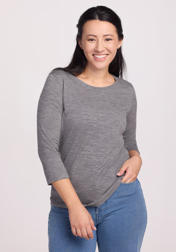 A woman with long dark hair, dressed in the Woolx Jenny 3/4 Sleeve Crew Neck in Graphite Heather and blue jeans, stands smiling confidently against a plain light background.