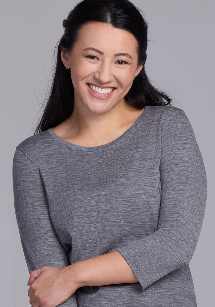 A woman with long dark hair smiles warmly, arms gently crossed, wearing the Woolx Jenny 3/4 Sleeve Crew Neck in Graphite Heather against a plain light gray background.