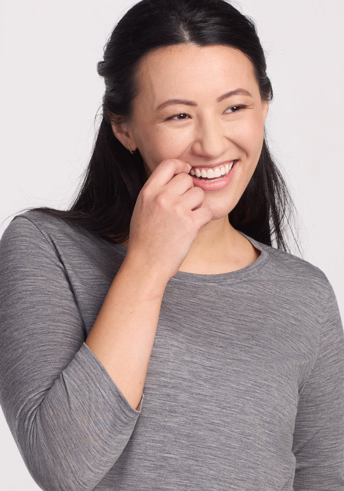 A woman with long dark hair wears the Woolx Jenny 3/4 Sleeve Crew Neck in Graphite Heather, smiling as she touches her face and looks slightly to the side against a plain light background.