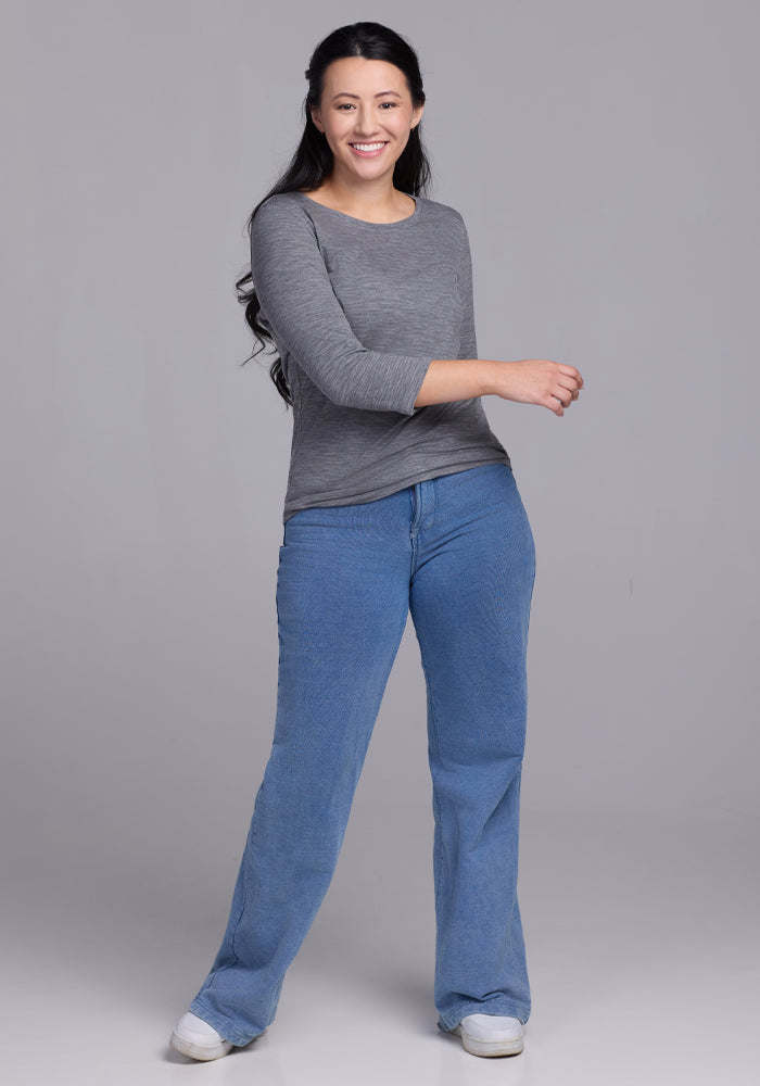 A woman with long dark hair, wearing a Woolx Jenny 3/4 Sleeve Crew Neck in Graphite Heather, blue jeans, and white sneakers smiles against a plain gray background.