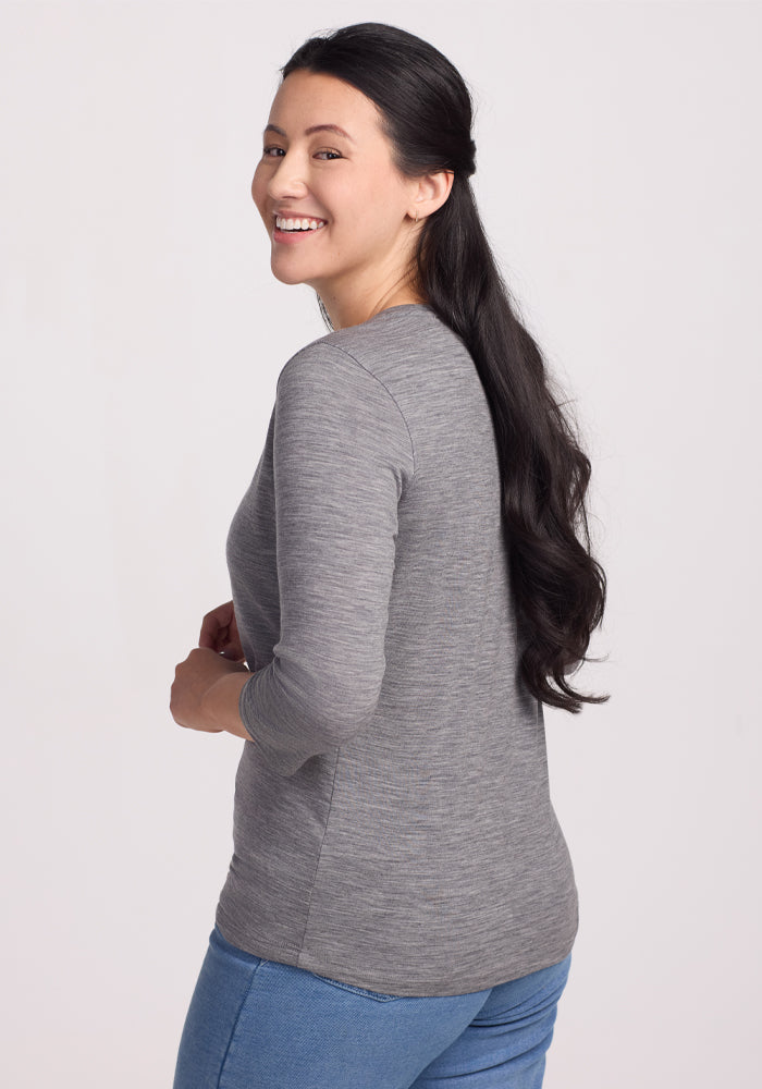 A woman with long dark hair, wearing the Woolx Jenny 3/4 Sleeve Crew Neck in Graphite Heather and blue jeans, smiles as she looks over her shoulder at the camera against a plain light background.