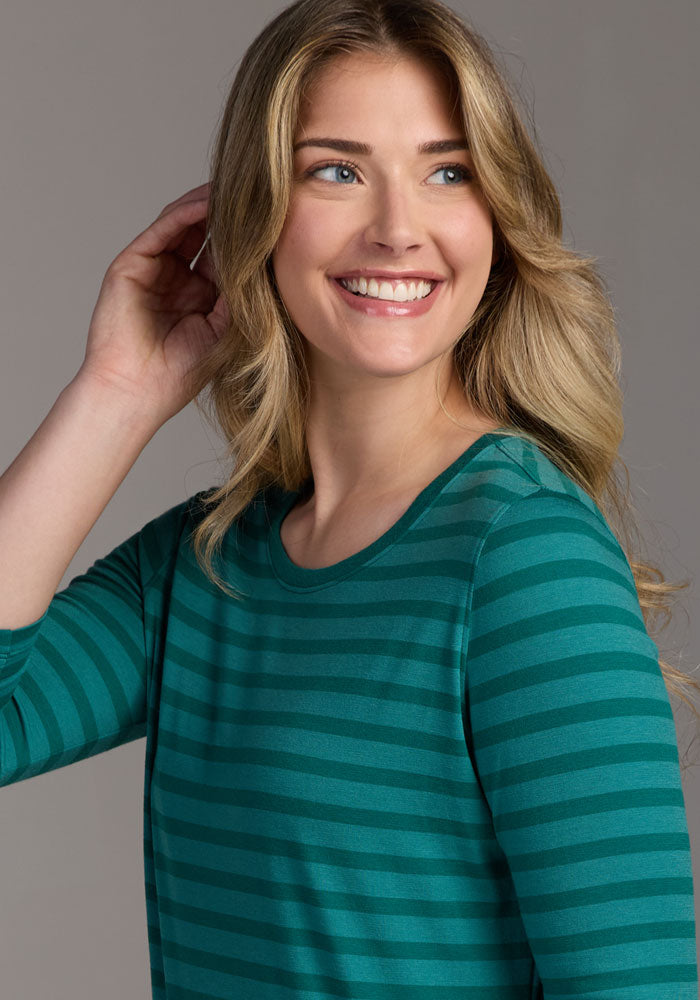 A woman with long, wavy blonde hair smiles and looks to the side, wearing the Woolx Jenny 3/4 Sleeve Crew Neck in Woodland Stripe. She holds one hand up near her head against a plain background.