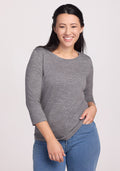 A woman with long dark hair, dressed in the Woolx Jenny 3/4 Sleeve Crew Neck in Graphite Heather and blue jeans, stands smiling confidently against a plain light background.