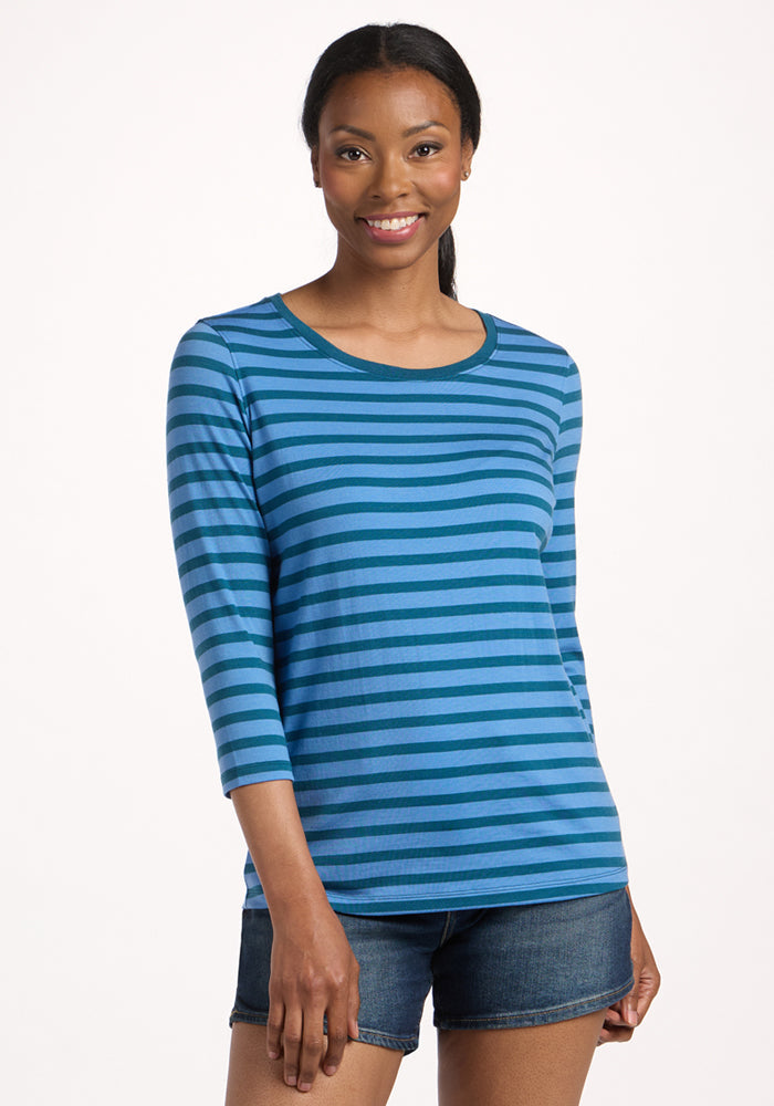 Smiling woman with long hair wearing the Woolx Jenny 3/4 Sleeve Crew Neck in Coastal Stripe and denim shorts, standing against a plain light background. | Jade is 5’10”, wearing a size S