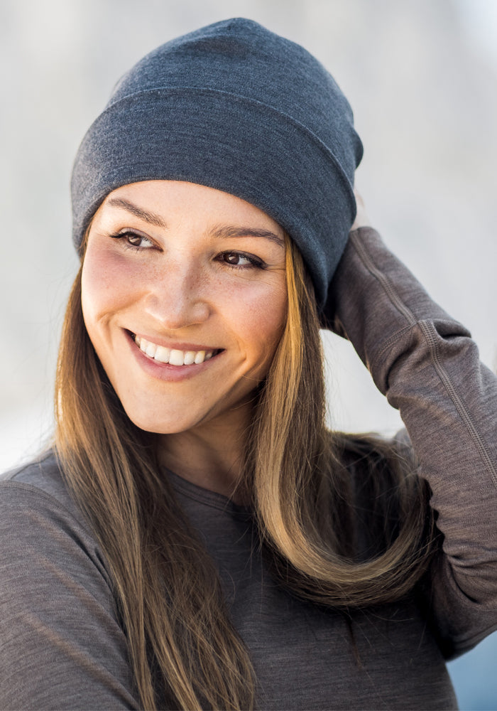 A woman with long brown hair smiles, looking to the side with one hand touching her Woolx Journey Hat in Winter Moss. She wears a matching brown long-sleeve shirt against a softly blurred Australian Merino wool background.