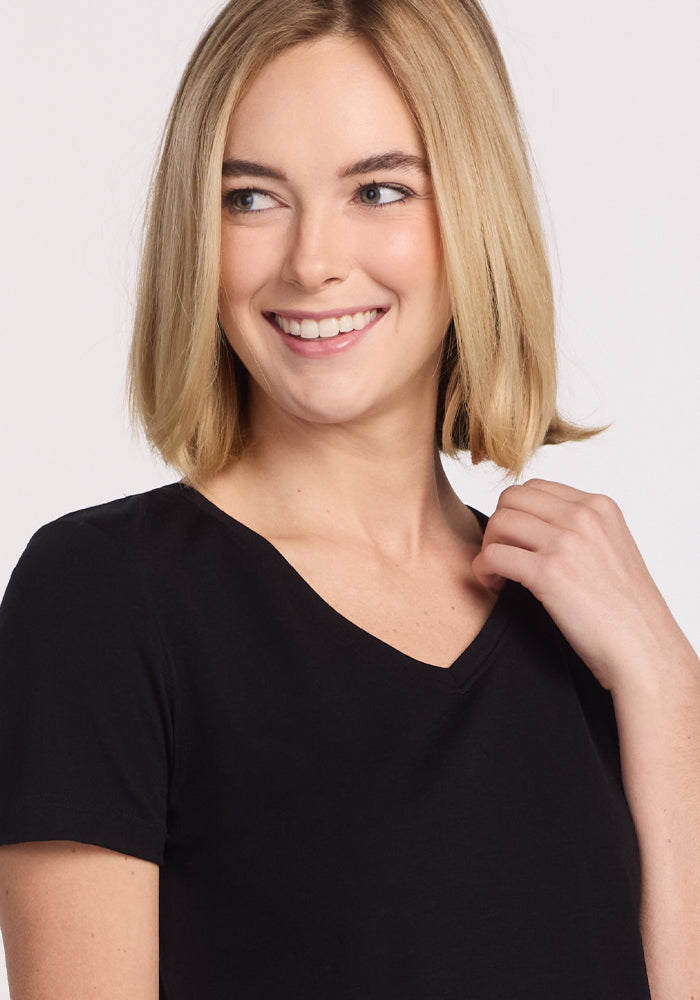 A young woman with straight blonde hair smiles and looks left, touching her collar as she wears the Woolx Junie V Neck - Black. Its relaxed fit and odor-resistant Merino wool fabric complete her stylish, comfortable look.