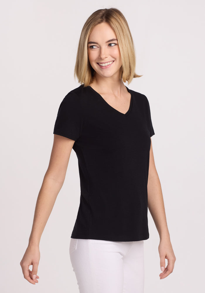 A woman with straight blonde hair, wearing the Woolx Junie V Neck in black and white pants, smiles while standing against a plain light background.