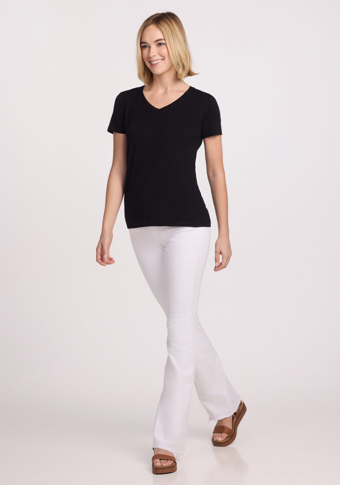 A smiling person with a short blonde bob is walking against a white background, wearing the Woolx Junie V Neck in black, white flared pants, and brown platform sandals.