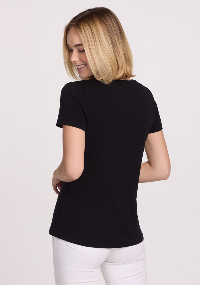A blonde woman, smiling and looking over her shoulder, wears the Woolx Junie V Neck in black and white pants. She stands facing away against a plain, light background. The tee is odor-resistant Merino wool.