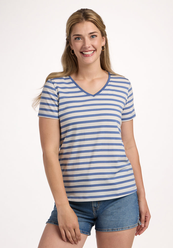 A smiling woman with long, light brown hair wears the Woolx Junie V Neck tee in Blue Sand Stripe and denim shorts, standing against a plain white background. | Ariana is 5'11", wearing a size S