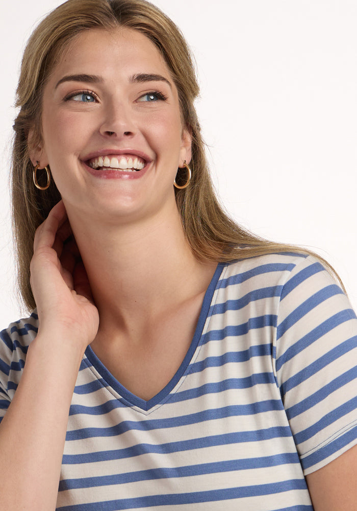 A woman with long light brown hair and gold hoop earrings smiles, touching her neck as she wears the odor-resistant Woolx Junie V Neck in Blue Sand Stripe against a plain white background.