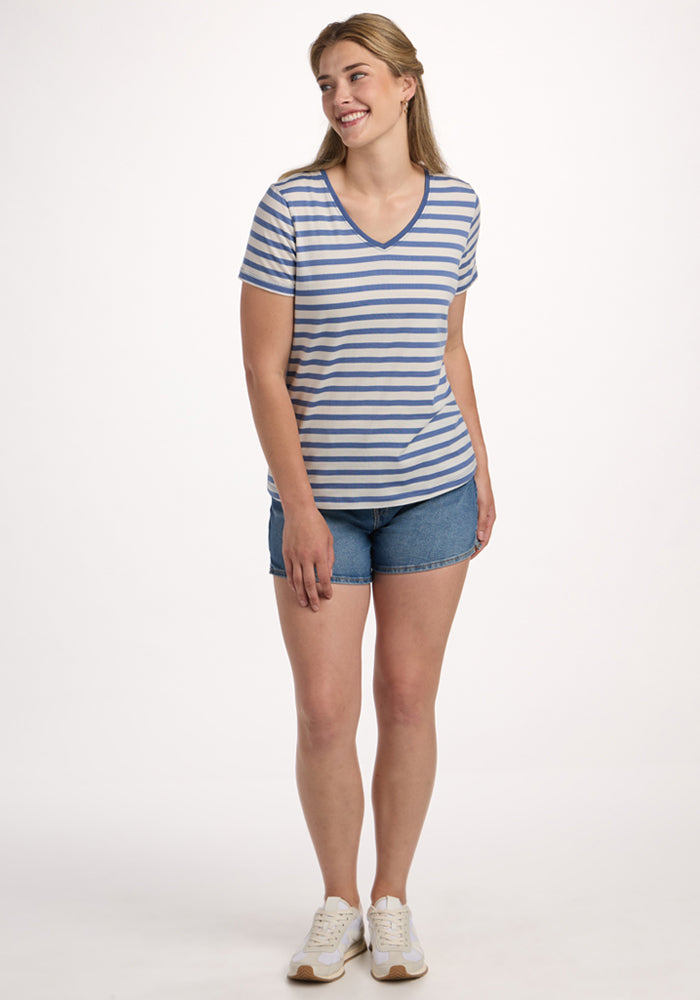 A woman smiles in the Woolx Junie V Neck - Blue Sand Stripe, paired with denim shorts and white sneakers, against a plain white background.