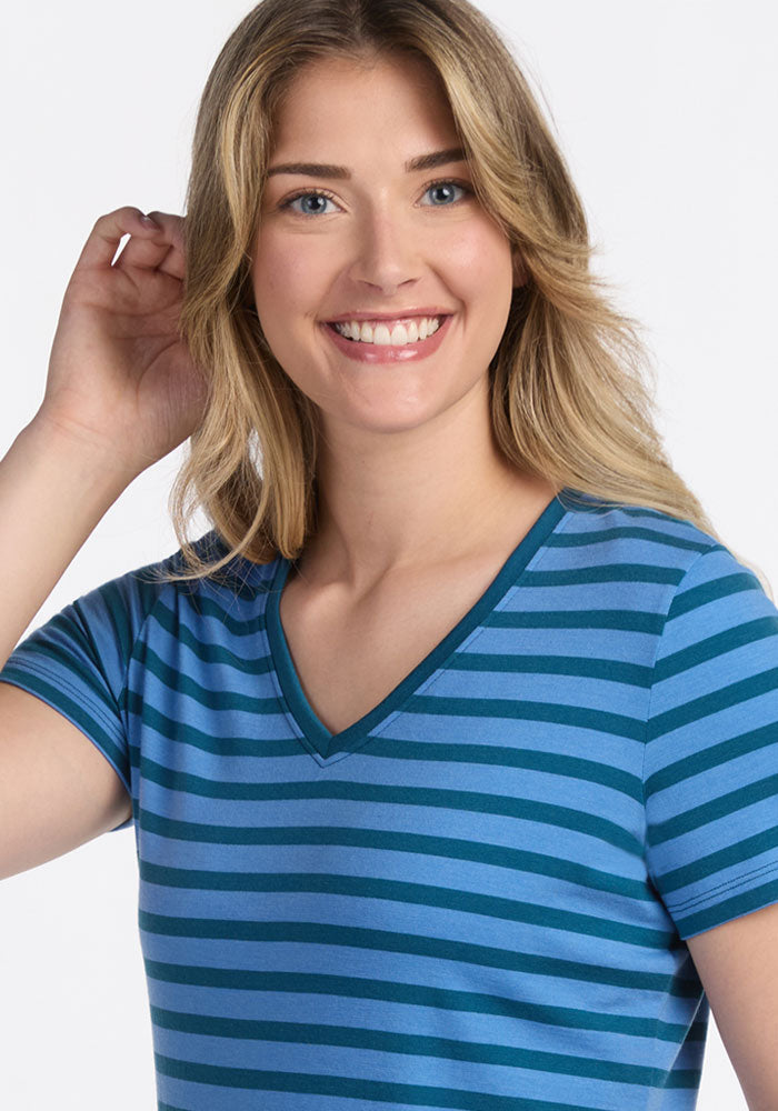 A smiling woman with long blonde hair wears the Woolx Junie V Neck - Coastal Stripe, a relaxed fit women's blue and teal striped short-sleeve tee, standing against a plain white background with one hand raised near her ear.