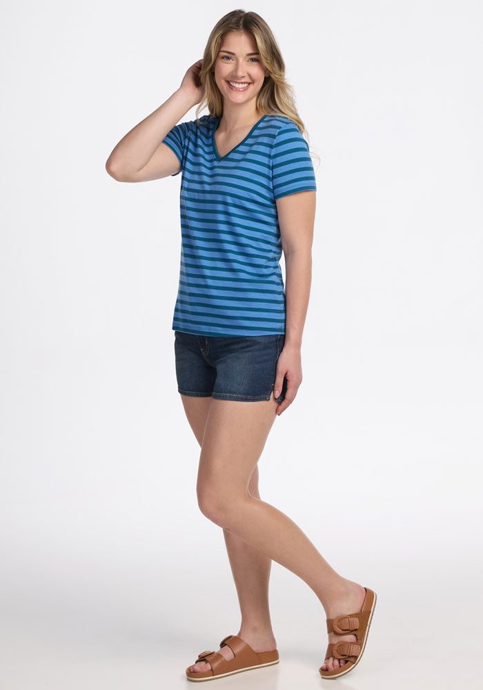 A smiling woman poses on one leg, wearing the Woolx Junie V Neck - Coastal Stripe shirt with dark denim shorts and brown sandals. She has long, wavy blonde hair and stands against a white background.