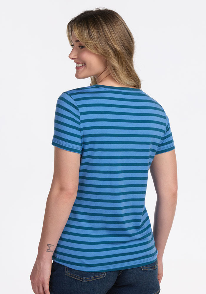 A woman with light brown hair smiles, standing away from the camera in Woolx's Junie V Neck - Coastal Stripe shirt and jeans. She has a small forearm tattoo. The background is plain white.