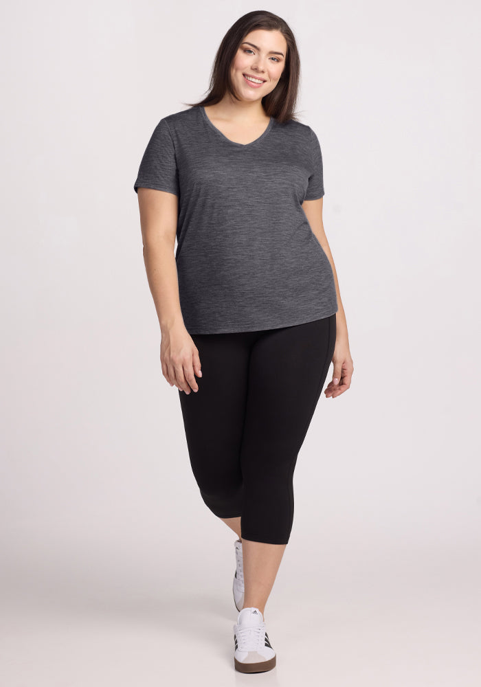 Woman in a Woolx Junie V Neck tee, cinder melange, with black leggings and white sneakers stands smiling.