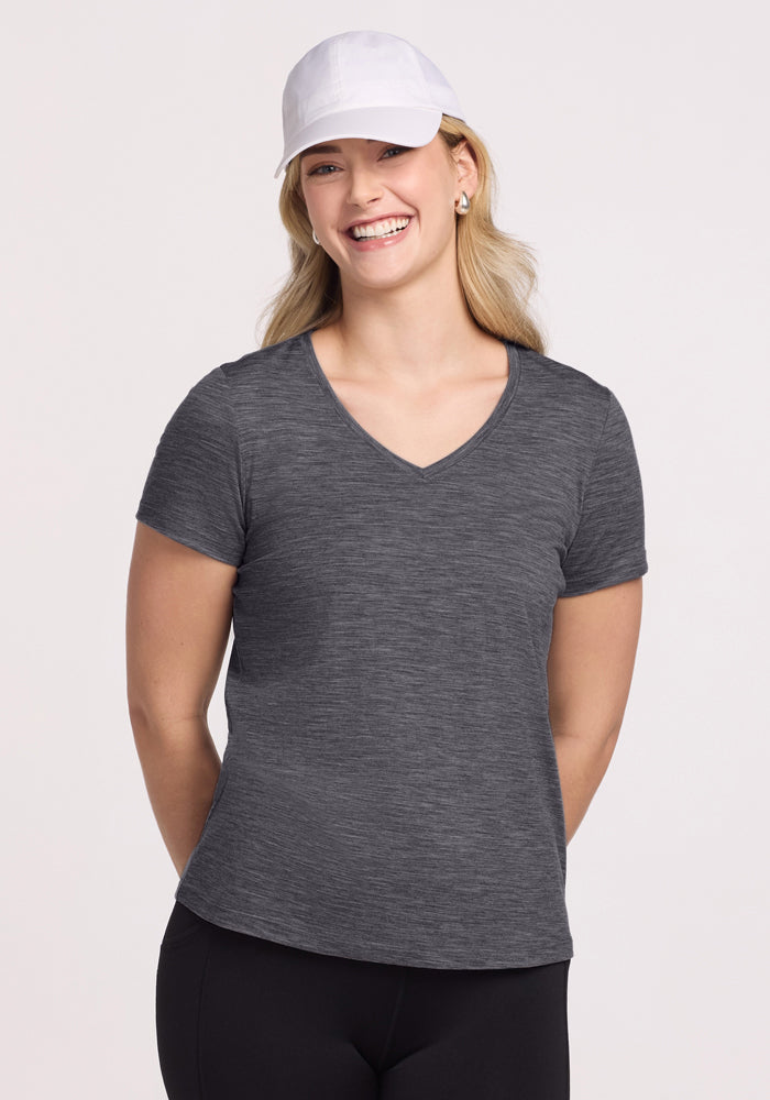 Smiling woman wears Woolx Junie V Neck in Cinder Melange, black pants, and a white cap against a plain background.