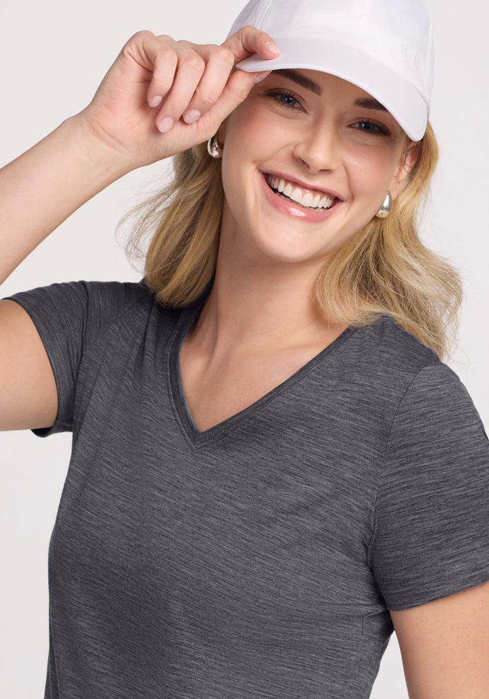 Smiling woman in a Woolx Junie V Neck (Cinder Melange) tee and white cap, holding her hat’s brim.