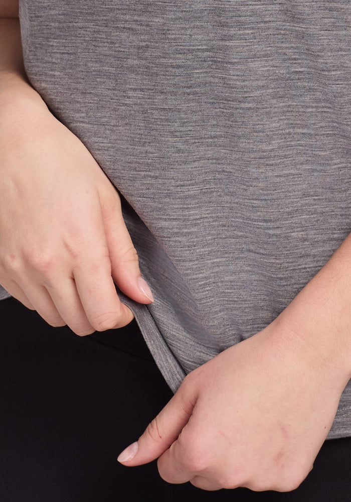 Close-up of a person wearing the Woolx Junie V Neck in Graphite Heather and black pants, gently pulling at the hem of the shirt with both hands.