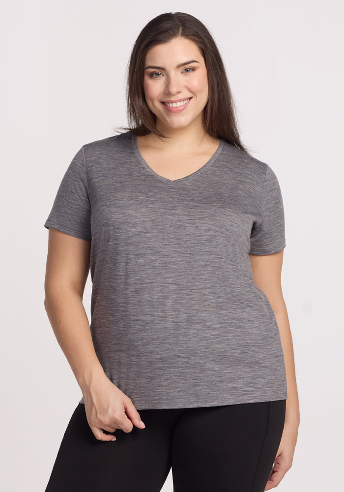 A woman with long brown hair smiles at the camera, wearing the Woolx Junie V Neck in Graphite Heather and black pants, against a plain light background. | Juliana is 5'10", wearing a size XL