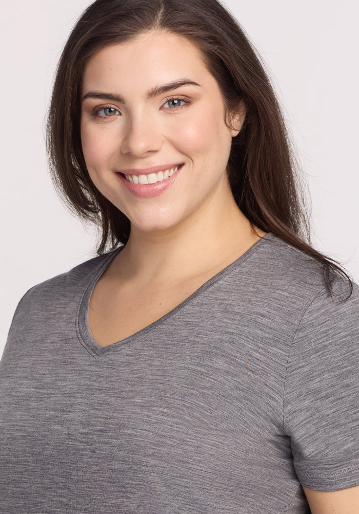 A woman with long brown hair smiles at the camera, wearing the Woolx Junie V Neck in Graphite Heather—an ultra-soft Merino wool tee with a relaxed fit, known for moisture-wicking comfort—posed against a plain light background.