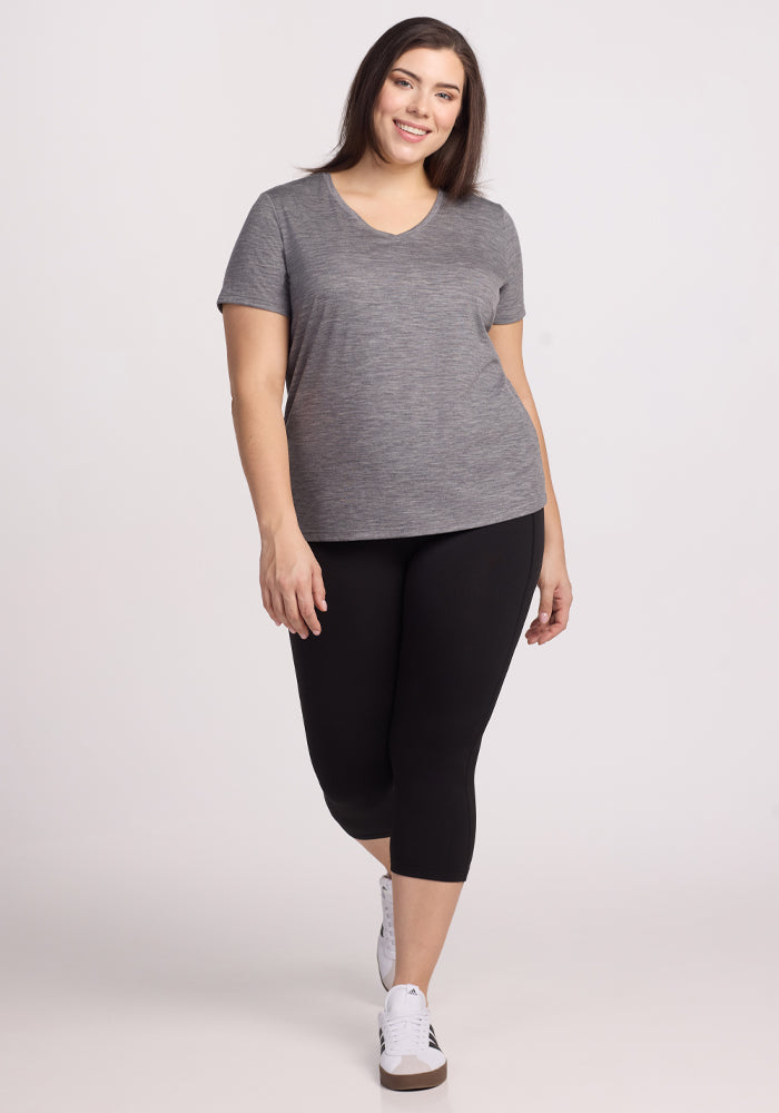 A woman with long brown hair is smiling and walking confidently in a Woolx Junie V Neck - Graphite Heather tee, black capri leggings, and white sneakers against a plain light-colored background.