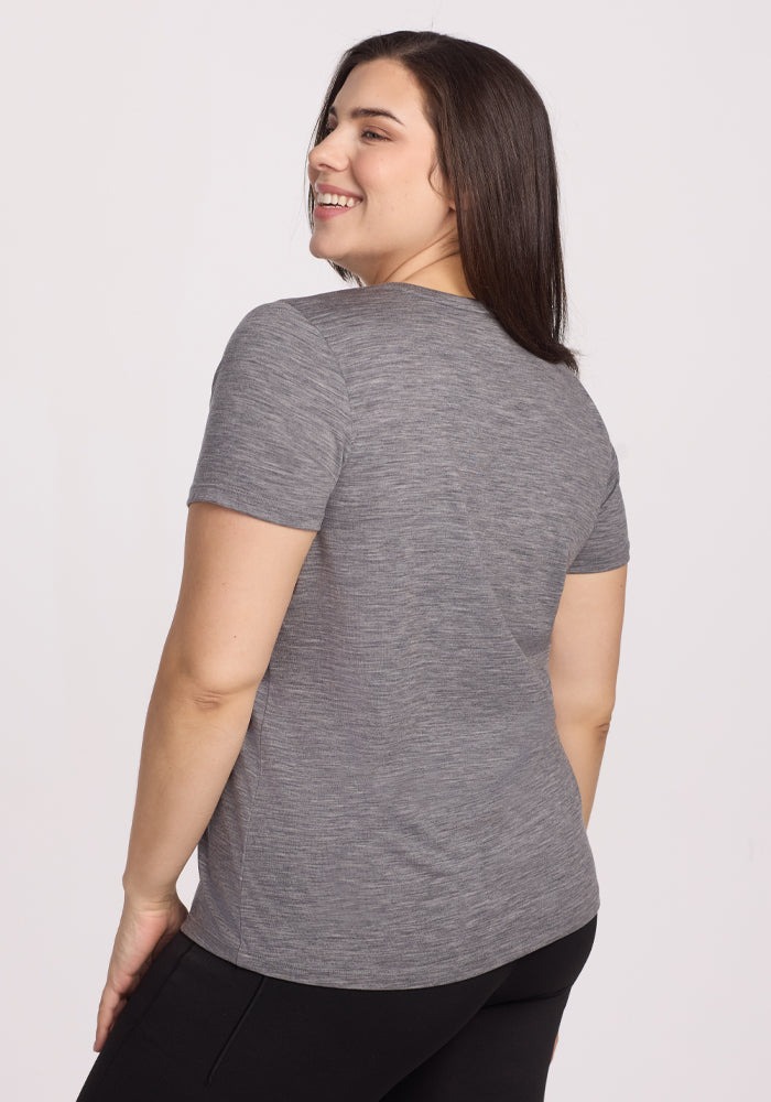 A woman with long dark hair smiles while looking over her shoulder, wearing the Woolx Junie V Neck in Graphite Heather and black pants, photographed from the back against a light background.