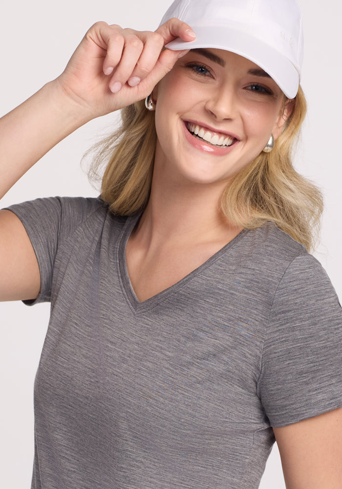 A smiling woman with light hair faces the camera against a plain background, wearing the Woolx Junie V Neck in Graphite Heather and a white cap, tilting its brim with one hand.