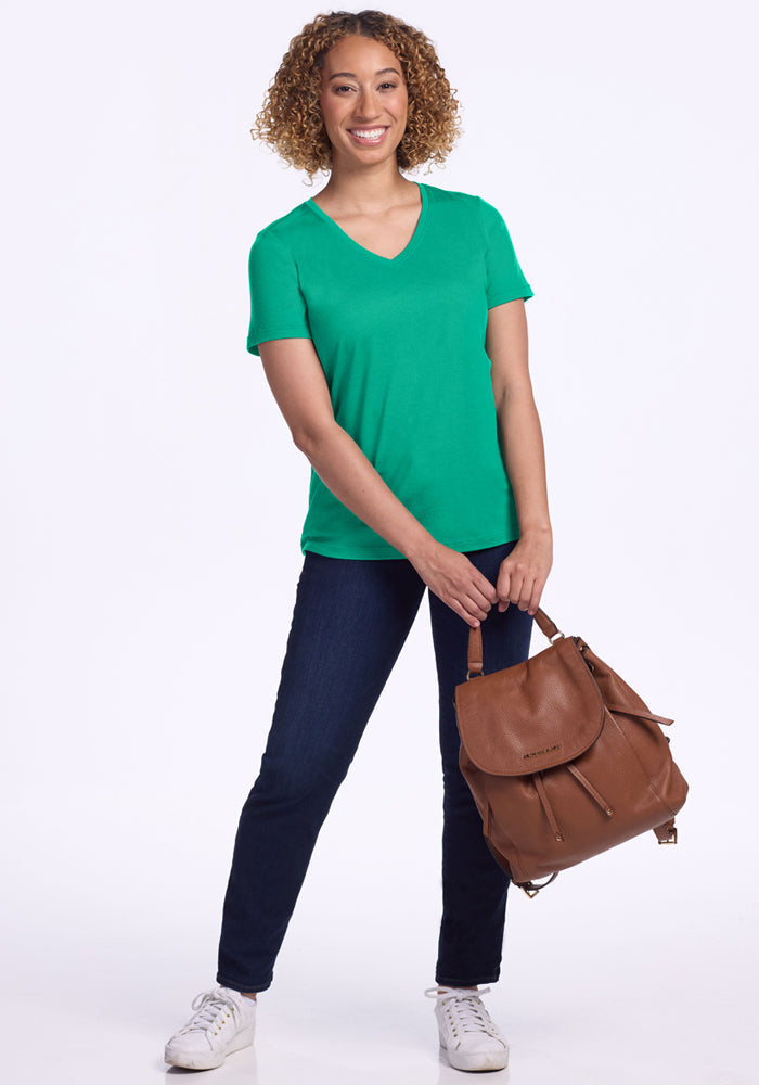 Woman wearing Woolx Junie V Neck - Jungle, dark jeans, white sneakers, holding a brown backpack, smiling.