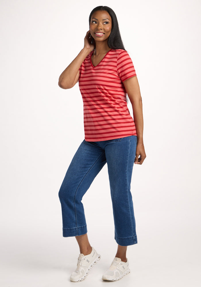 A woman smiles in the Woolx Junie V Neck - Wildberry Stripe, featuring red and pink stripes, styled with blue cropped jeans and white sneakers, posed against a plain white background.
