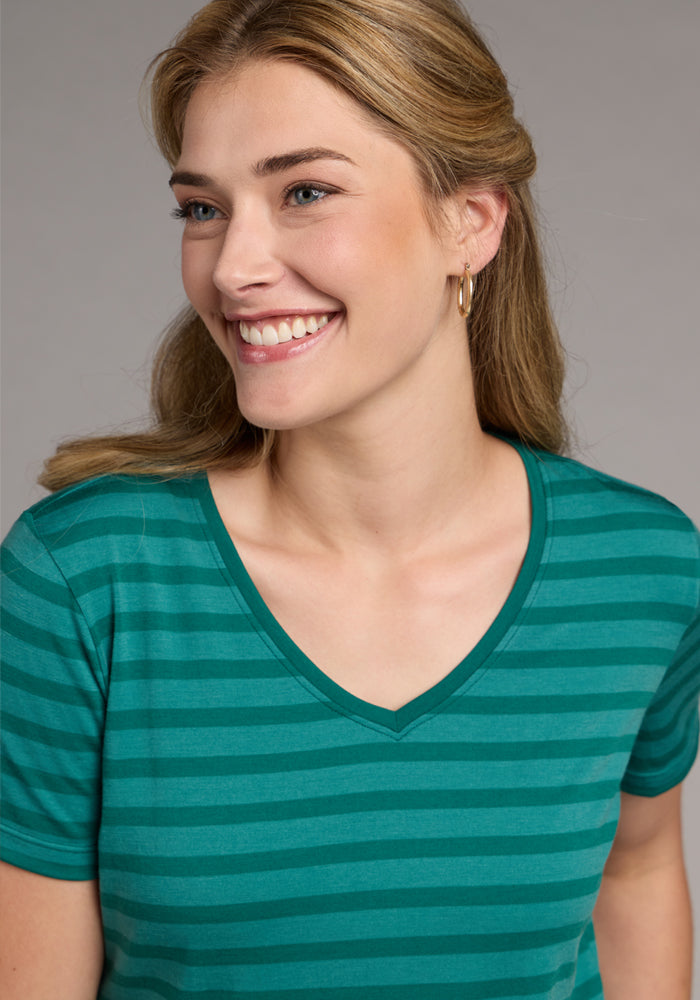 A smiling woman with light brown hair wears the Woolx Junie V Neck - Woodland Stripe tee and small hoop earrings, standing against a neutral background.