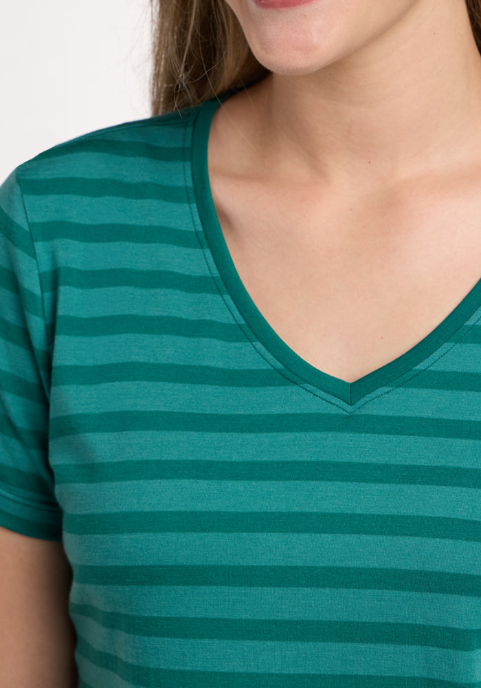 A close-up shows a woman in the Woolx Junie V Neck - Woodland Stripe, featuring green and teal stripes. She is smiling slightly, with only her lower face, neck, and upper torso visible.