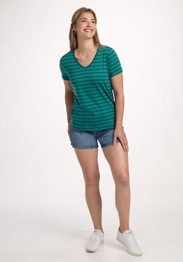 A woman smiles against a white background wearing the Woolx Junie V Neck in Woodland Stripe, blue denim shorts, and white sneakers. Her hair is pulled back, one hand on her thigh, creating a casual, comfortable look.