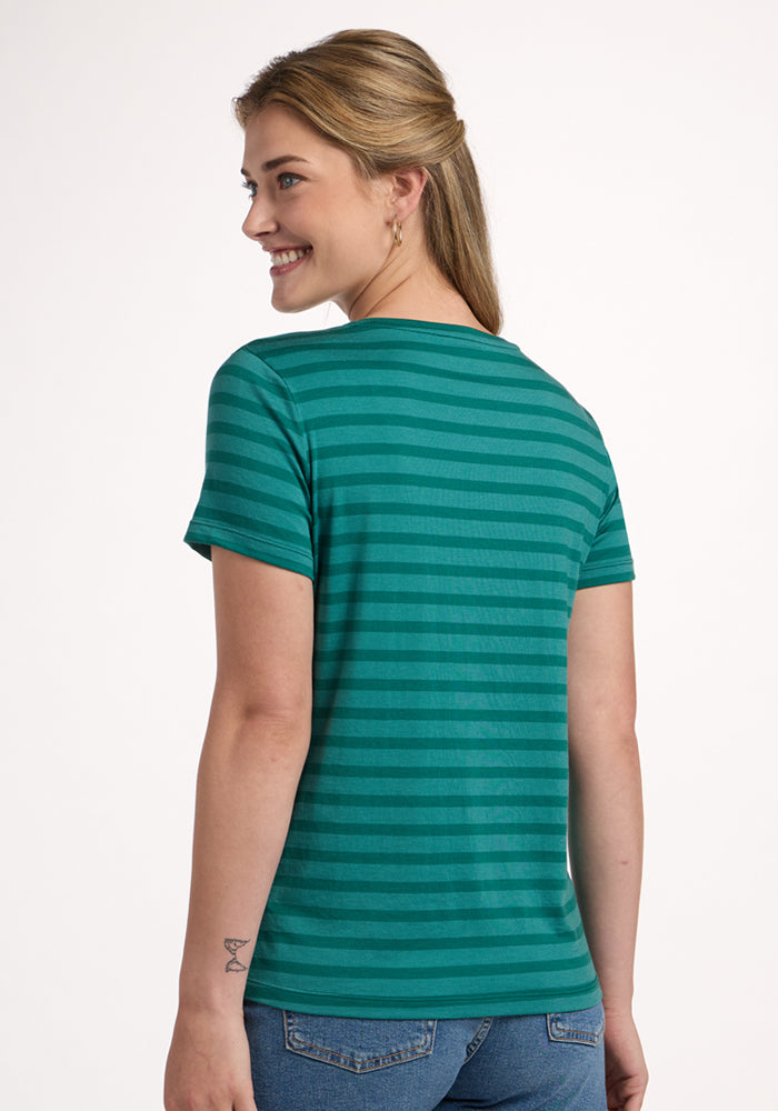 A blonde woman smiles over her shoulder, wearing the Woolx Junie V Neck in Woodland Stripe and blue jeans. The relaxed fit tee stands out against a light background, showing off her butterfly tattoo on her left wrist.