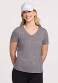 A woman with long blonde hair, in a white cap and a relaxed-fit Woolx Junie V Neck tee in Graphite Heather that's odor-resistant, smiles with her hands behind her back against a plain light background.