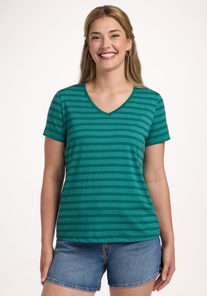 A woman smiles in the Woolx Junie V Neck – Woodland Stripe, a relaxed fit green and teal striped tee, paired with denim shorts against a plain white background. | Ariana is 5'11", wearing a size S