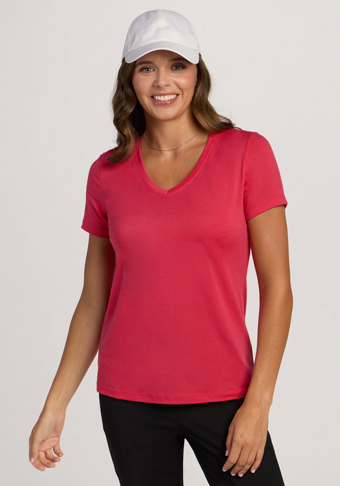 Smiling woman in Woolx Junie V Neck - Berry Ice Pop, black pants, and white cap stands against a light background. | Madeleine is 5’10”, wearing a size XS