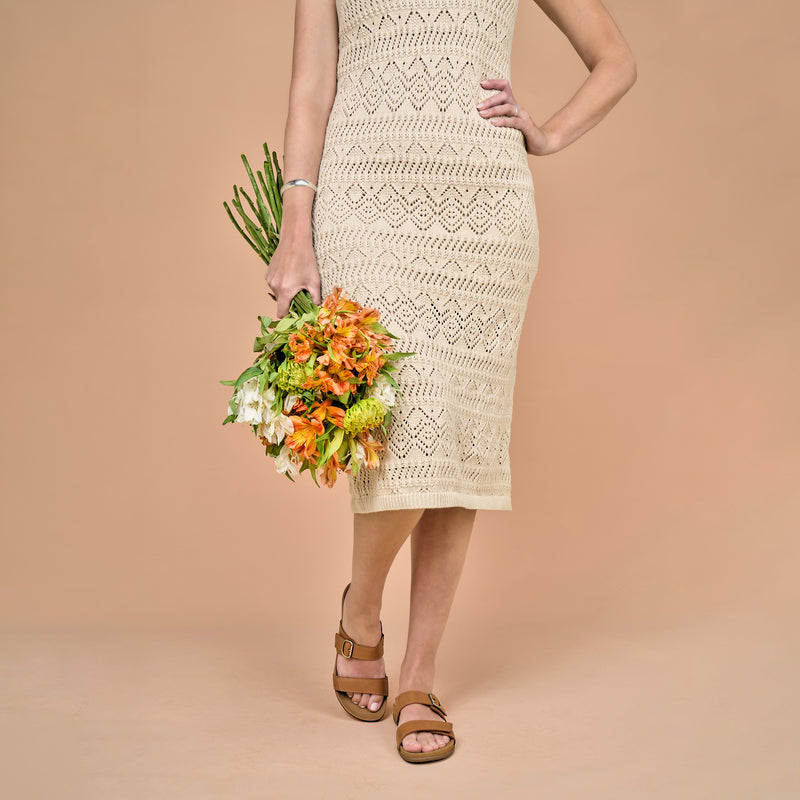 A woman in a dress holding flowers and wearing brown leather slide sandals.