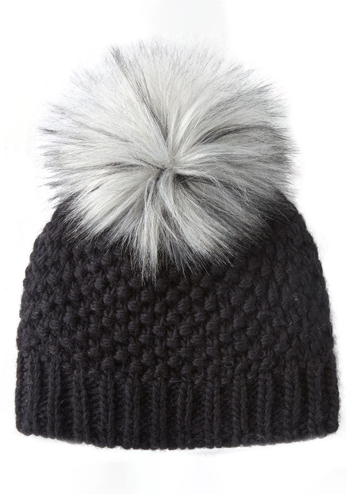 The Kaylee Beanie OLD by Woolx, a black, chunky knit hat made from soft Merino wool, is adorned with a large, fluffy faux fur pom-pom on top and is set against a plain white background.