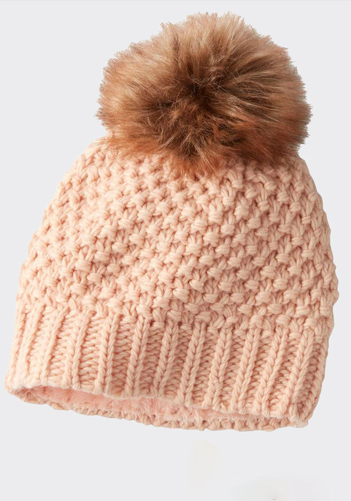 The Woolx Kaylee Beanie OLD is a chunky knit hat in pink, crafted from Merino wool and showcasing a textured pattern. It is adorned with a large, fluffy faux fur pom-pom on top and is set against a plain backdrop.
