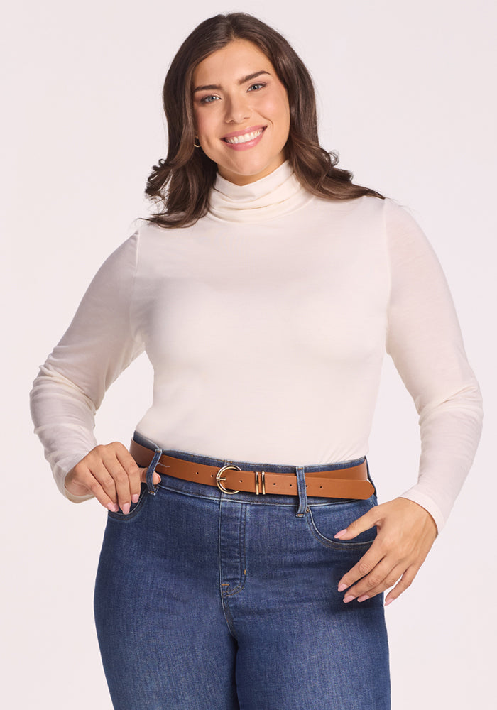 A smiling woman with long brown hair wears the Woolx Kennedy Bodysuit in Buttercream, paired with blue jeans and a brown belt. She stands against a plain light background, hands near her waist, highlighting this versatile layering essential. | Juliana is 5'10", wearing a size XL