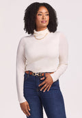 Wearing the Woolx Kennedy Bodysuit in Buttercream, a woman with curly hair pairs it with blue jeans, a chain necklace, and a belt as she stands against a plain light background, gazing slightly to the side with a gentle smile. | Maysa is 5’3”, wearing a size M