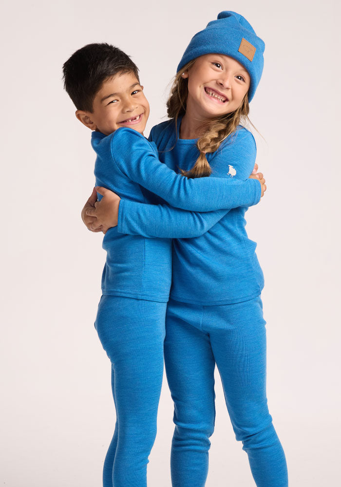 Two young children in matching blue thermal outfits smile and hug. One wears the Woolx Kids Baylor Beanie in Blue Razz, standing out against a plain light background that highlights their cozy style.